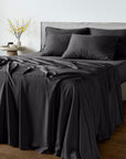 A neatly made bed with bambaw cozy black bamboo bedding, including a sheet, pillowcases, and a draped throw blanket, set in a modern bedroom with a gray upholstered headboard, wooden floor, and a small side table with yellow flowers.