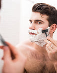 Man shaving his face with a Bambaw safety razor and shaving cream in front of a mirror.