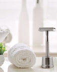 Bambaw Silver safety razor with stand on a white surface with a rolled-up towel and plant in the background