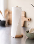 A roll of reusable white paper towels on a wooden stand on a kitchen counter, with a green bowl and wooden cutting boards blurred in the background.