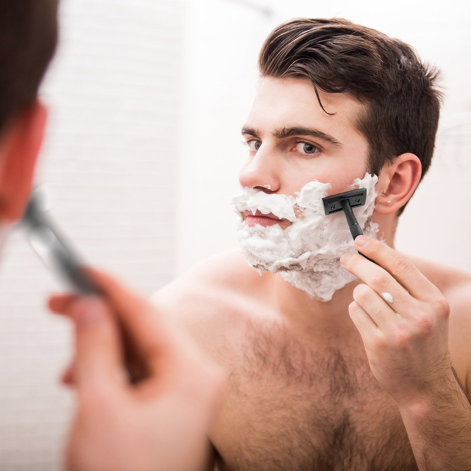 Man shaving his face with a bambaw black metal safety razor and shaving cream in front of a mirror.