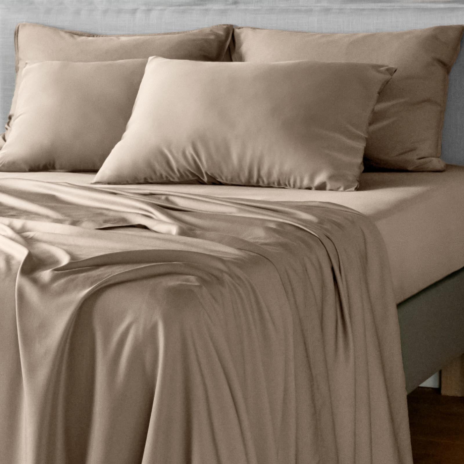 A neatly made bed with taupe-beige bamboo bedding, including a fitted sheet, matching pillowcases, and two large pillows, shown in a softly lit bedroom setting.