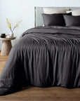 A bed dressed in black bambaw cozy bamboo duvet cover and matching pillow shams, styled in a bedroom with a light gray headboard, wood floor, and a small side table with a vase of yellow branches.