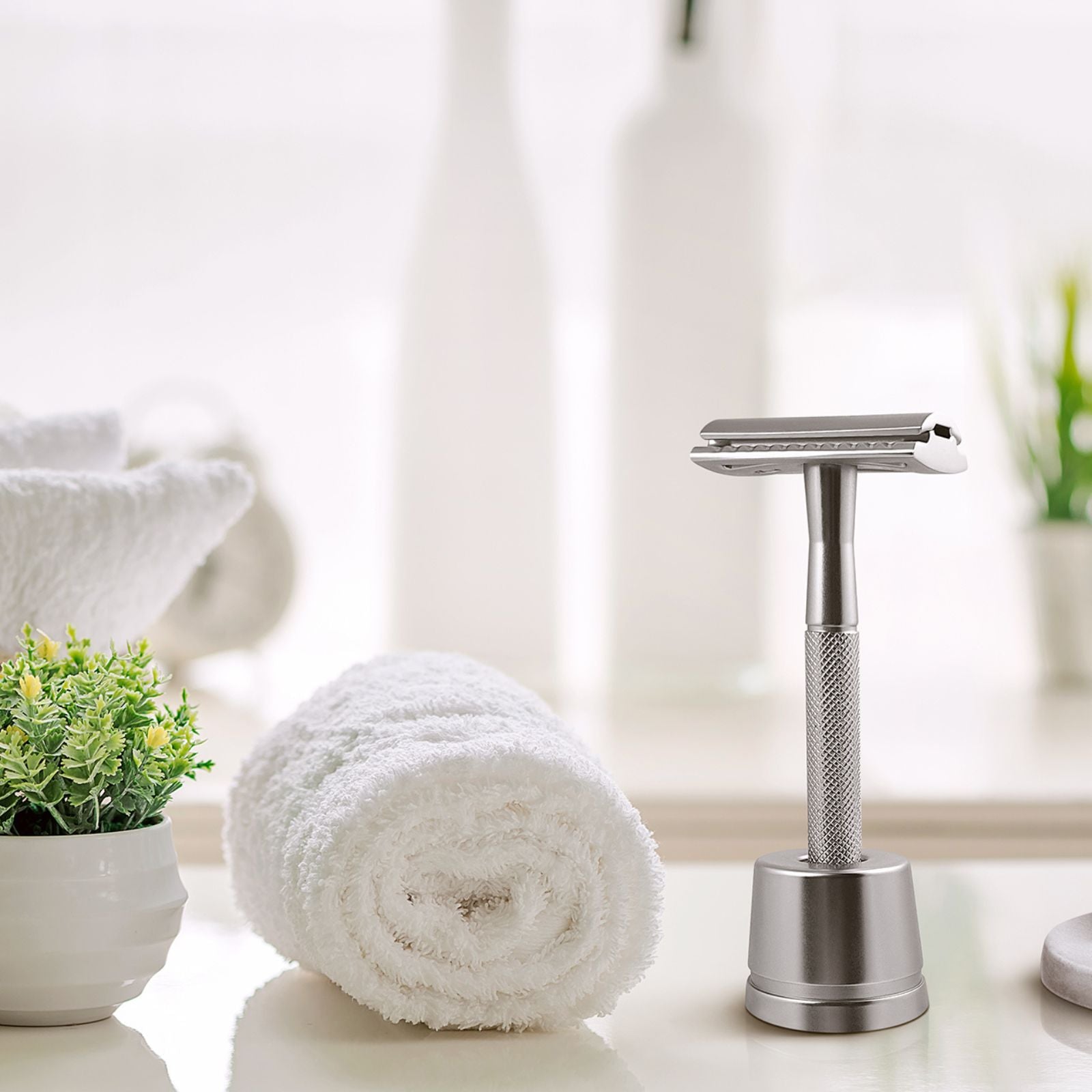 Bambaw Silver safety razor with stand on a white surface with a rolled-up towel and plant in the background