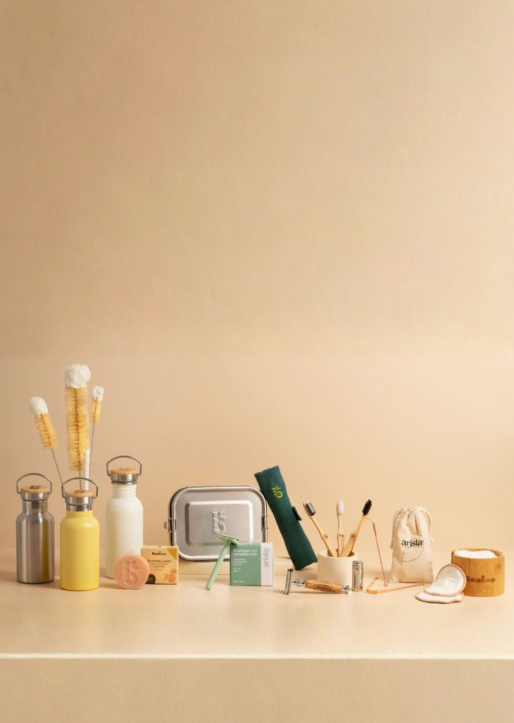 A curated collection of eco-friendly bathroom and travel accessories arranged on a beige surface against a matching beige background. The set includes stainless steel and reusable bottles, bamboo toothbrushes, a metal tin, a green rolled travel case, a small green box, cotton pads, and a bamboo candle.