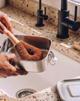 A plastic-free dish cleaning brush with a wooden handle is being held over a kitchen sink by two hands, while washing a stainless steel lunch box. The sink has a speckled terrazzo countertop, black faucet handles, and a bar of soap on a wooden block.