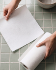 A close-up of hands unrolling a sheet of white reusable paper towel on a light green tiled surface.