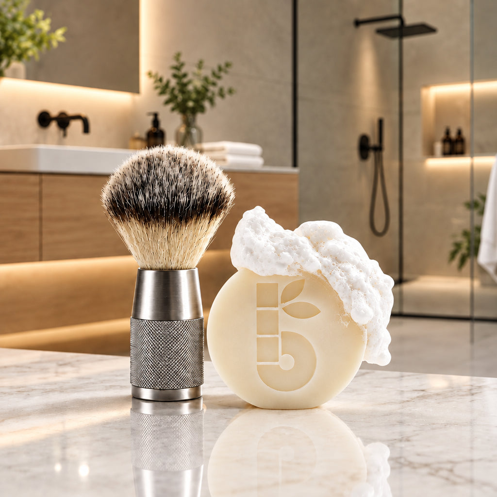 A bambaw metal shaving brush with dark and light vegan bristles and a textured silver handle stands beside a round cream-colored shaving soap bar with white lather on top, set on a marble countertop in a modern bathroom.