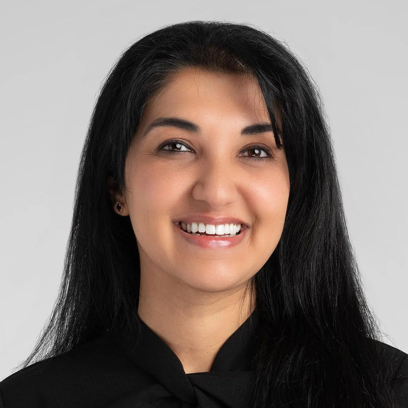 Dr Shipli-dermatologist. Woman with long black hair wearing a black outfit against a gray background